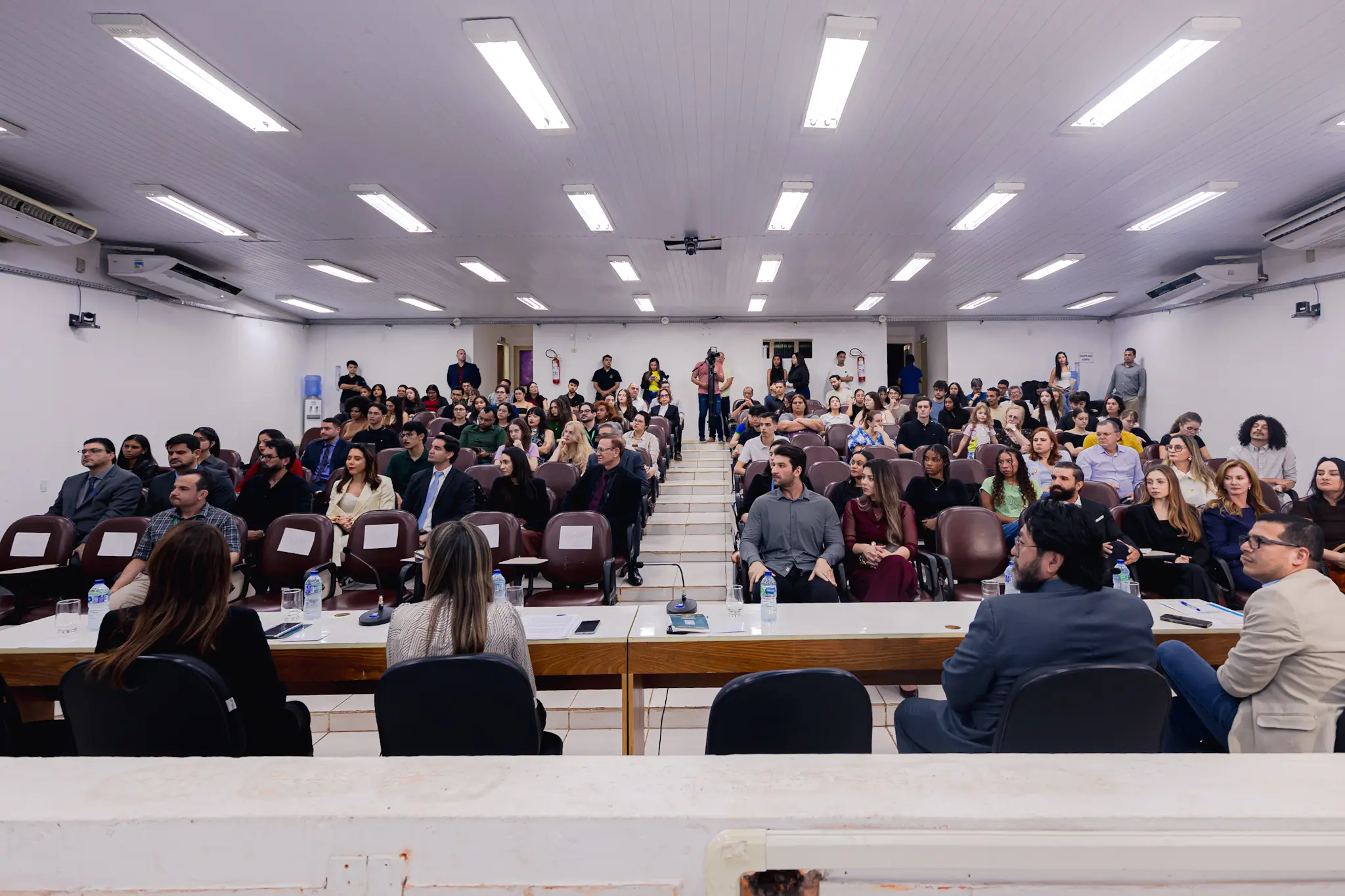 Vista de trás de uma mesa com quatro palestrantes de costas, voltados para uma plateia numerosa e sentada em um auditório iluminado com teto de PVC.