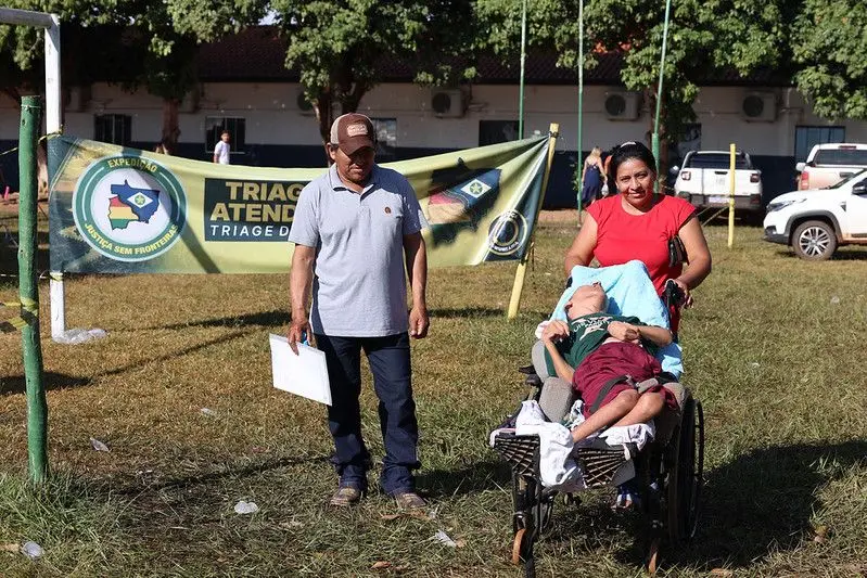 Homem e mulher conduzem uma pessoa em cadeira de rodas por uma área gramada. Ao fundo, há uma faixa de identificação da Expedição Justiça Sem Fronteiras e veículos estacionados.