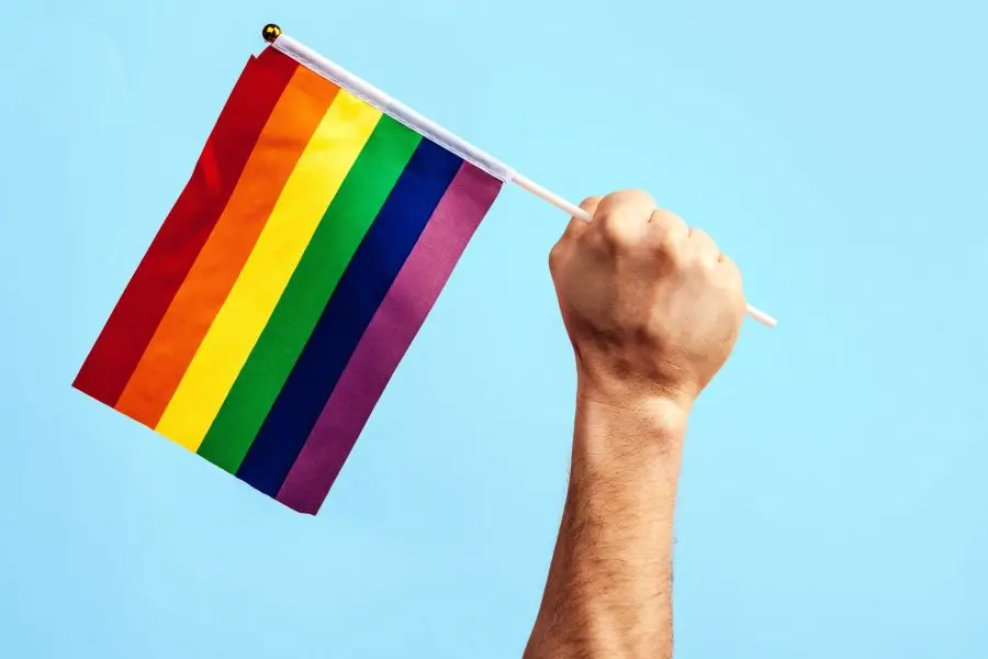 Mão ergue uma bandeira com as cores do arco íris contra um céu azul.