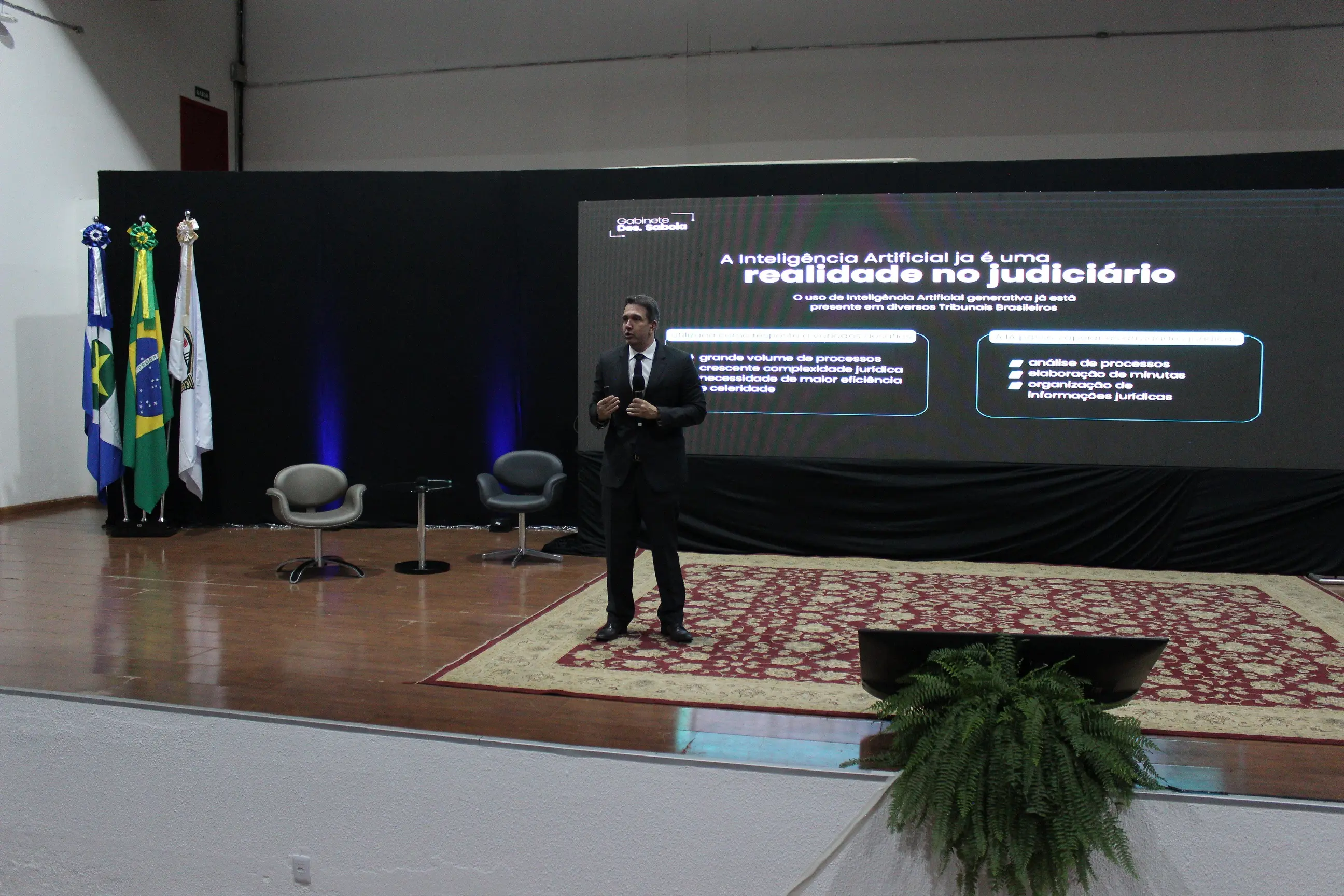 Homem de terno discursa em um palco de madeira sobre um tapete estampado. Atrás dele, um grande painel exibe um slide sobre Inteligência Artificial no Judiciário e há três bandeiras à esquerda.
