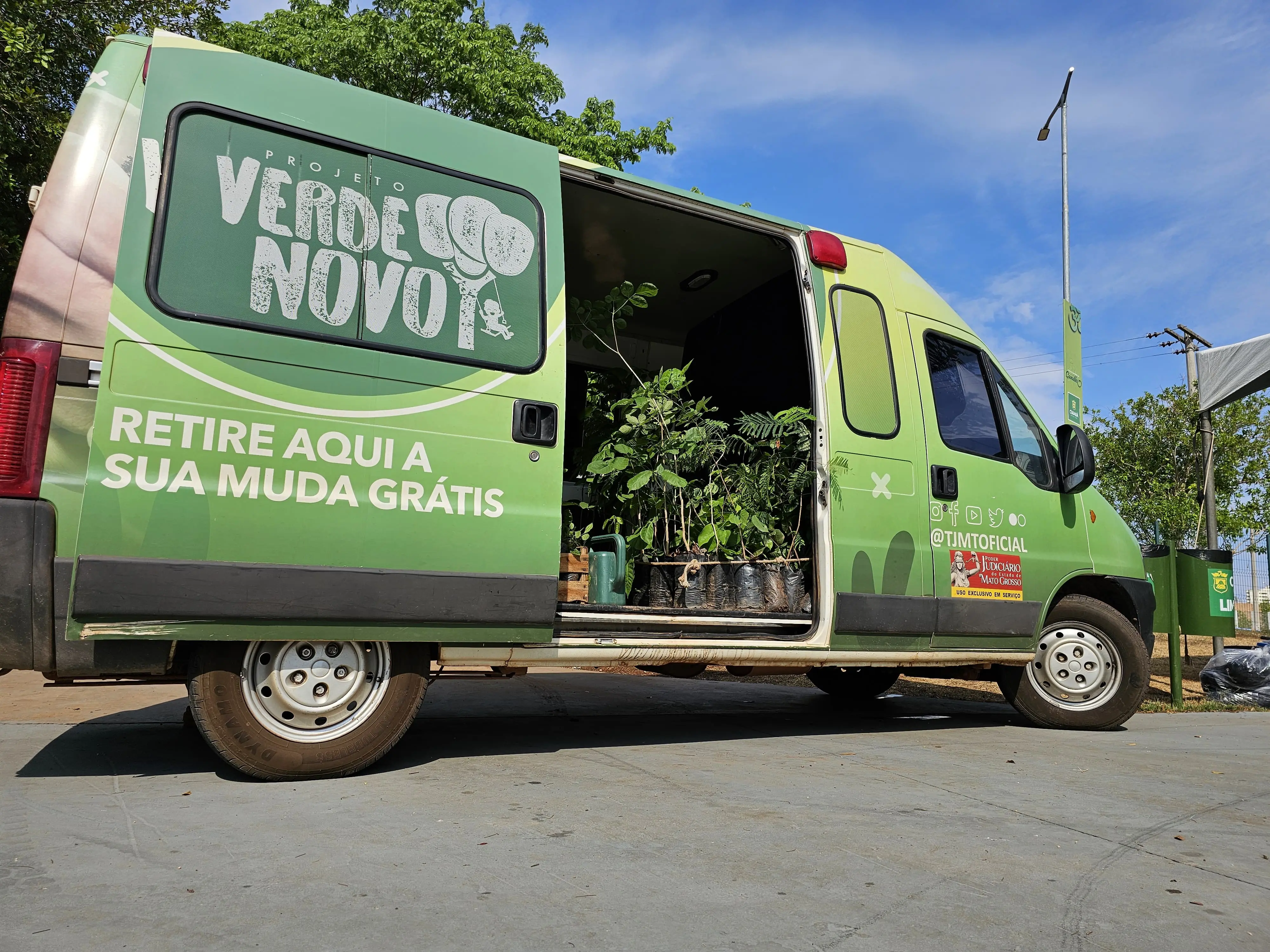 Van verde do Projeto Verde Novo está estacionada com a porta lateral aberta, exibindo diversas mudas de árvores. O veículo convida a população a retirar gratuitamente plantas para arborização urbana.