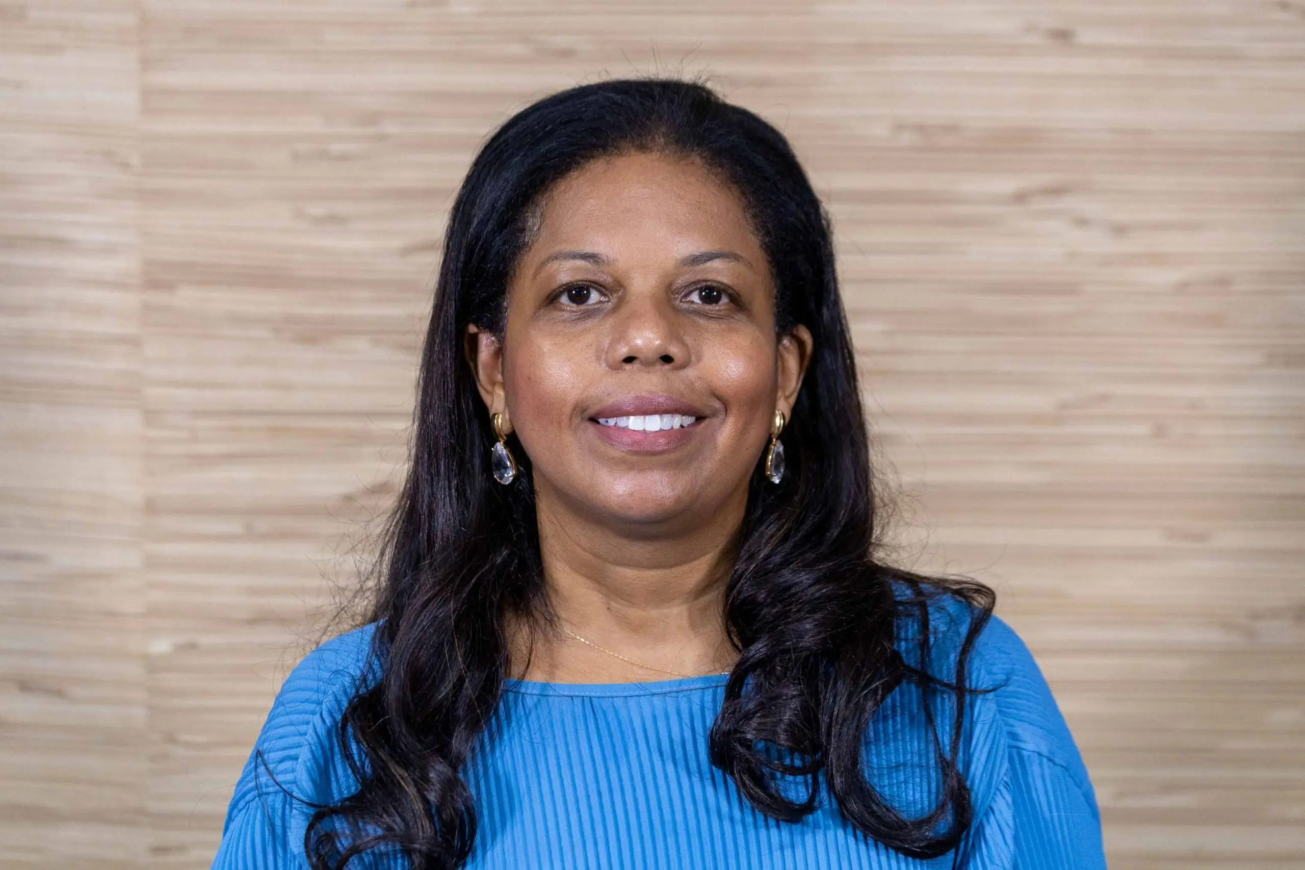 Foto horizontal que mostra a juíza Renata Evaristo sorrindo diretamente para a câmera. Ela é negra, tem longos cabelos escuros e ondulados soltos, e usa brincos pendentes de cristal e uma blusa azul. 