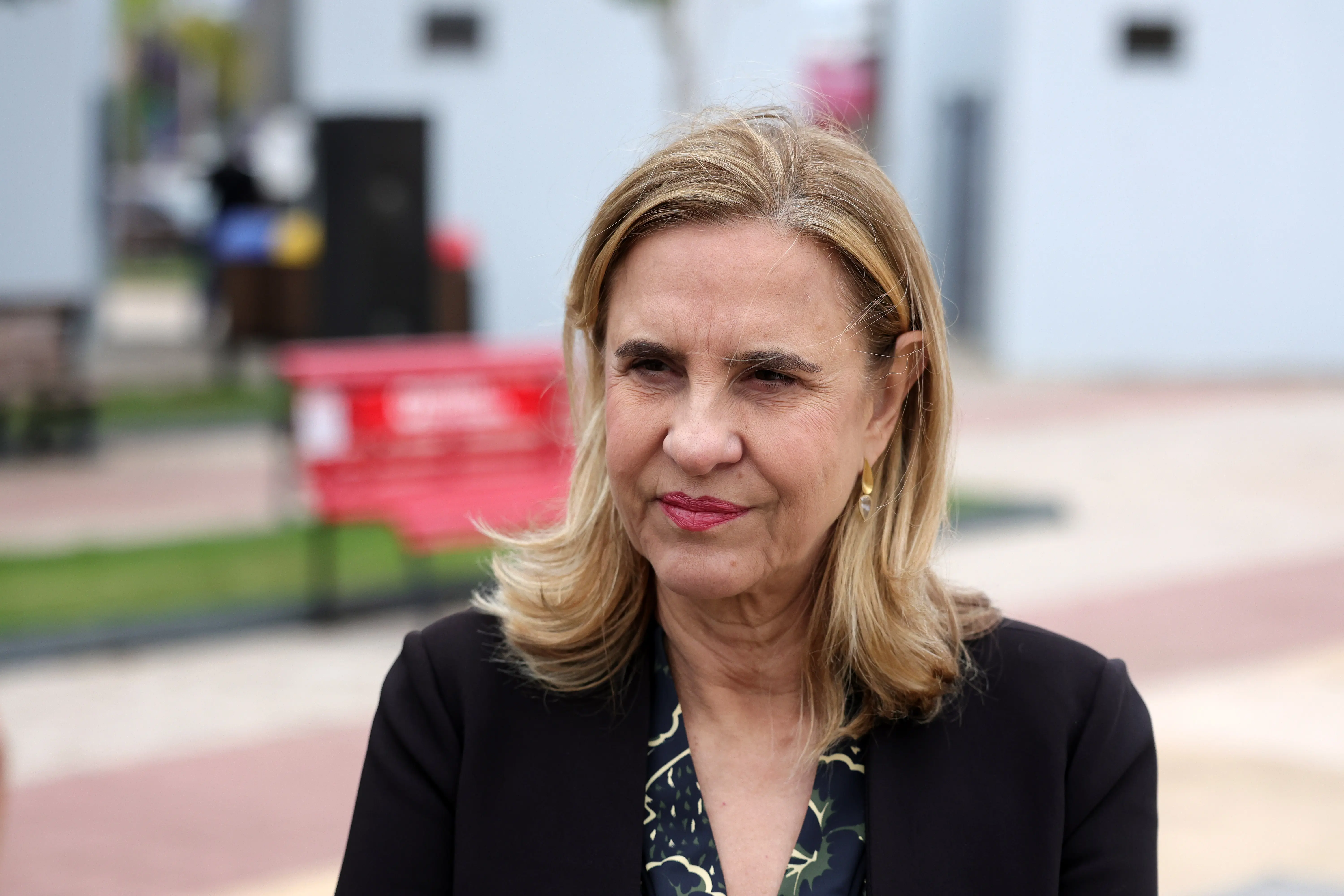 Retrato em primeiro plano de uma mulher loira de cabelos médios, vestindo blazer preto sobre blusa estampada. Ao fundo, um ambiente externo desfocado com a presença de um banco vermelho