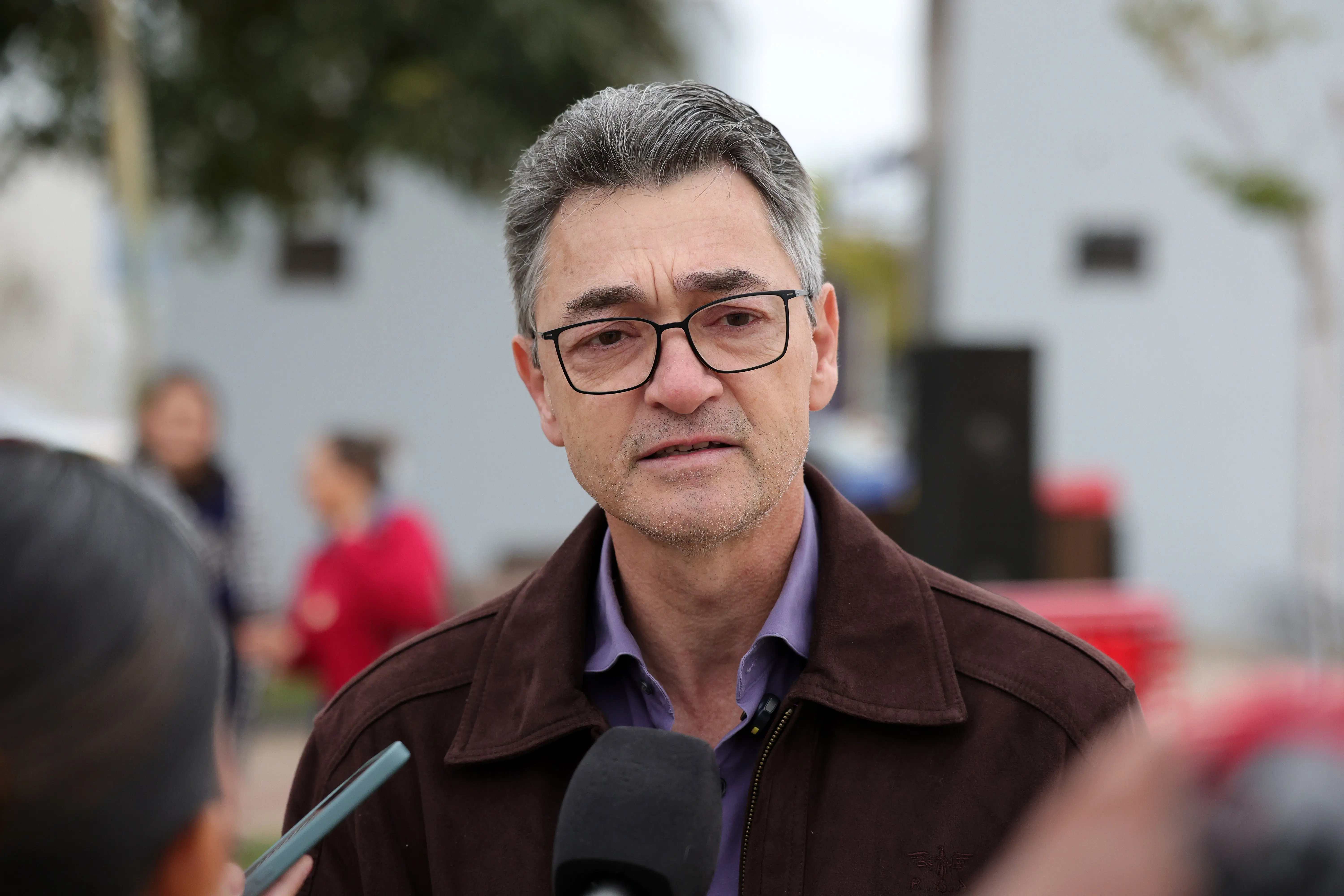 Homem de óculos, cabelos grisalhos, camisa roxa e jaqueta marrom concede entrevista em ambiente externo. Um microfone e um celular aparecem em primeiro plano. Fundo desfocado.
