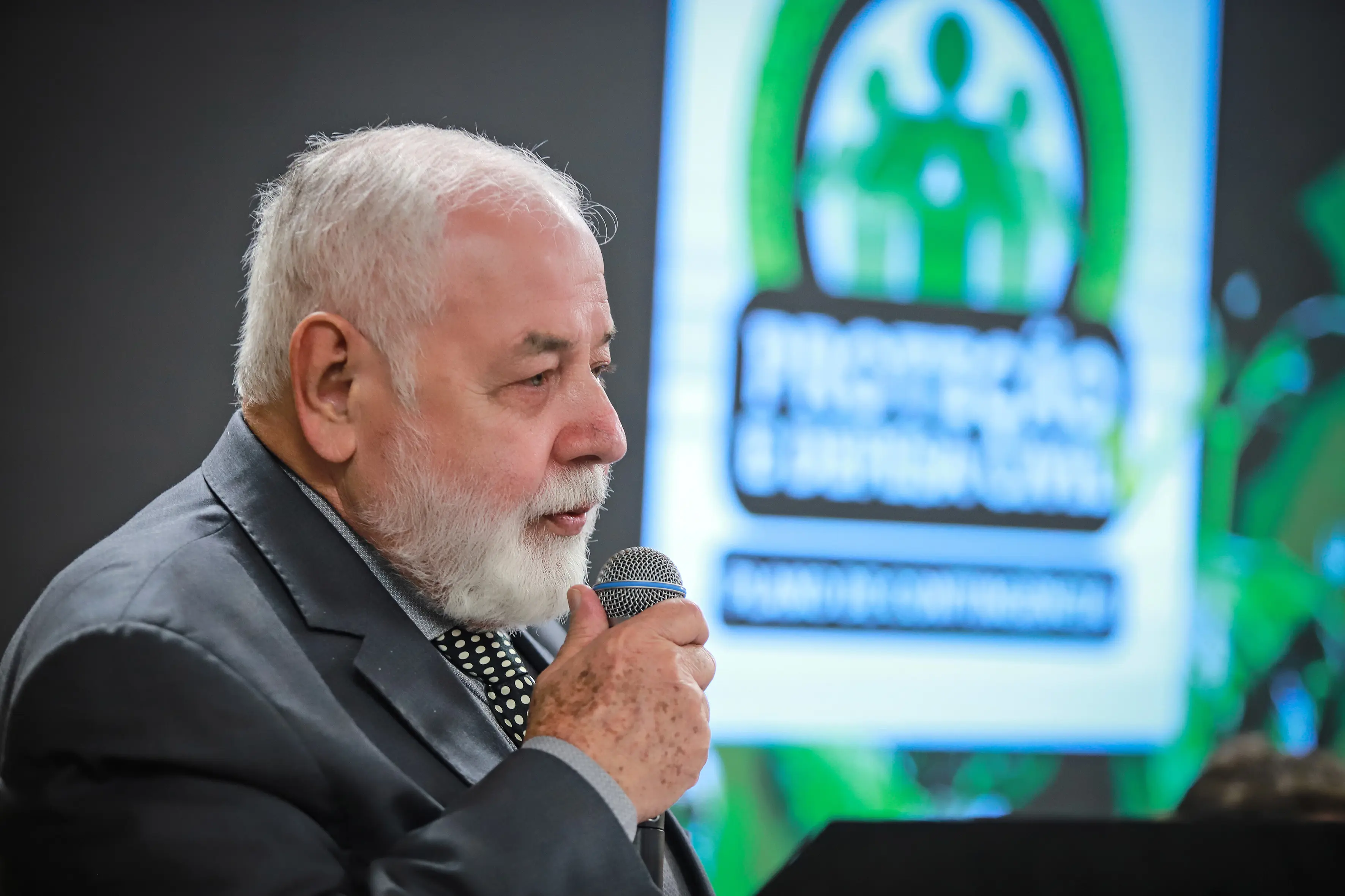Presidente do Poder Judiciário de Mato Grosso (TJMT), desembargador José Zuquim Nogueirade, um homem idoso, barba e cabelos brancos, de terno, com gravata de bolinhas, segurando um microfone e ao fundo um logo desfocado.