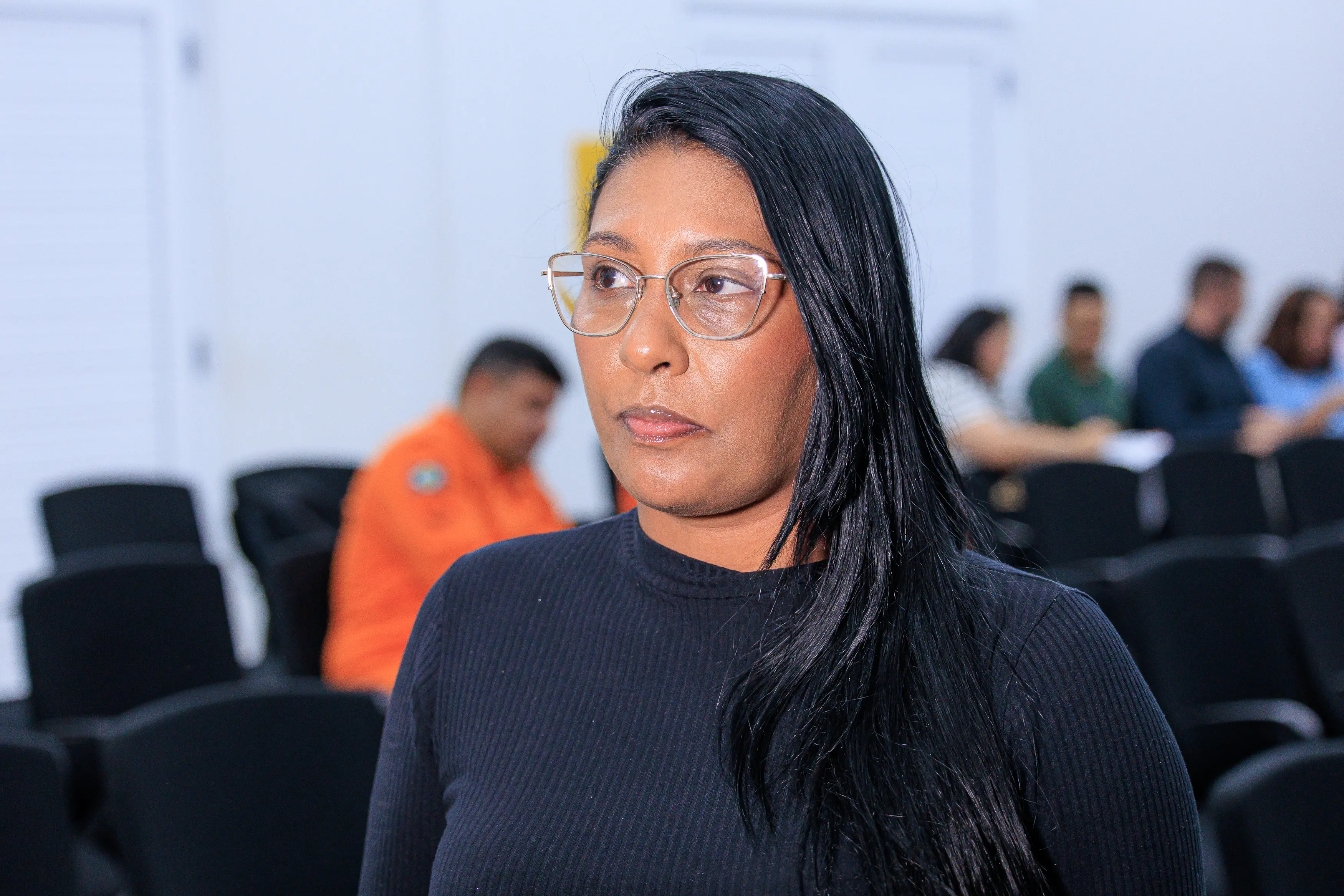Conselheira tutelar Eliane Mezari é uma mulher negra de cabelos longos e pretos e óculos com armação de metal dourado, olha para a direita. Ela veste uma blusa preta canelada de gola alta. O fundo é um auditório com cadeiras escuras e pessoas desfocadas.