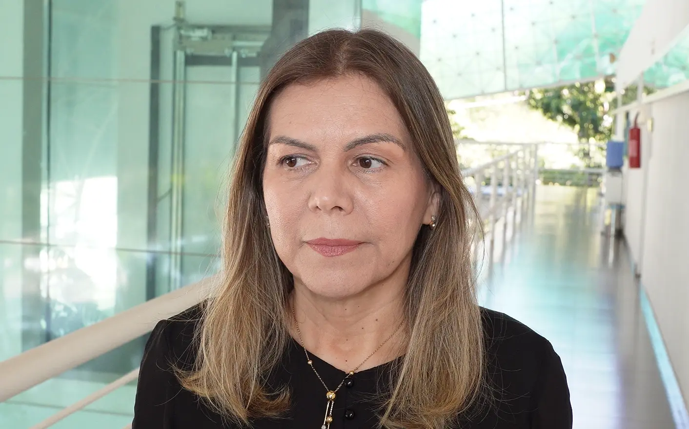 Promotora Lindinalva Correia é uma mulher de pele clara, cabelos castanhos médios lisos e soltos. Veste blusa preta, colar dourado e olha levemente para a esquerda. Ao fundo, um corredor interno com paredes de vidro e piso claro.