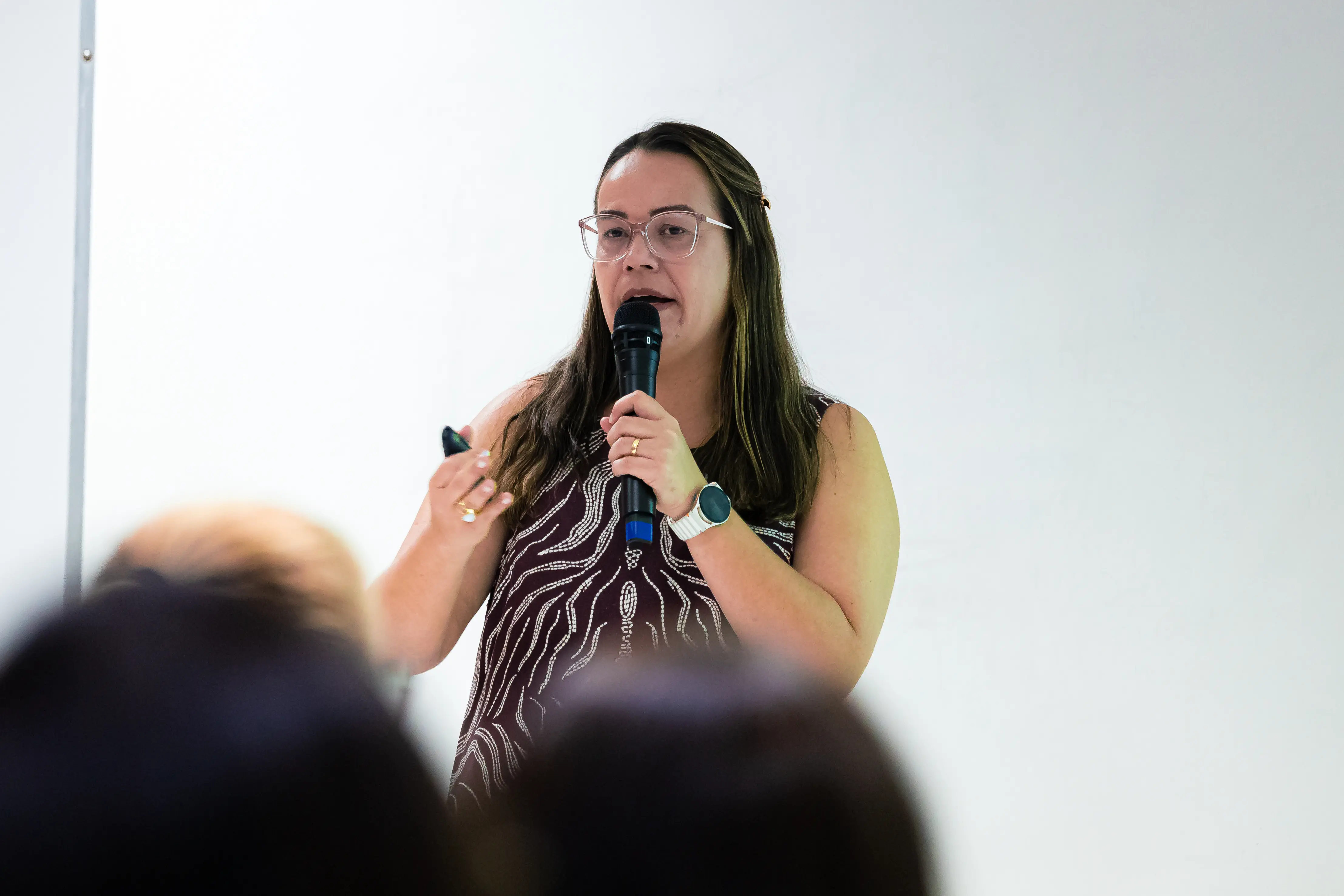 Mulher de óculos e vestido estampado fala ao microfone, segurando um passador de slides. Ela palestra diante de uma plateia, cujas cabeças aparecem desfocadas em primeiro plano. Fundo branco.
