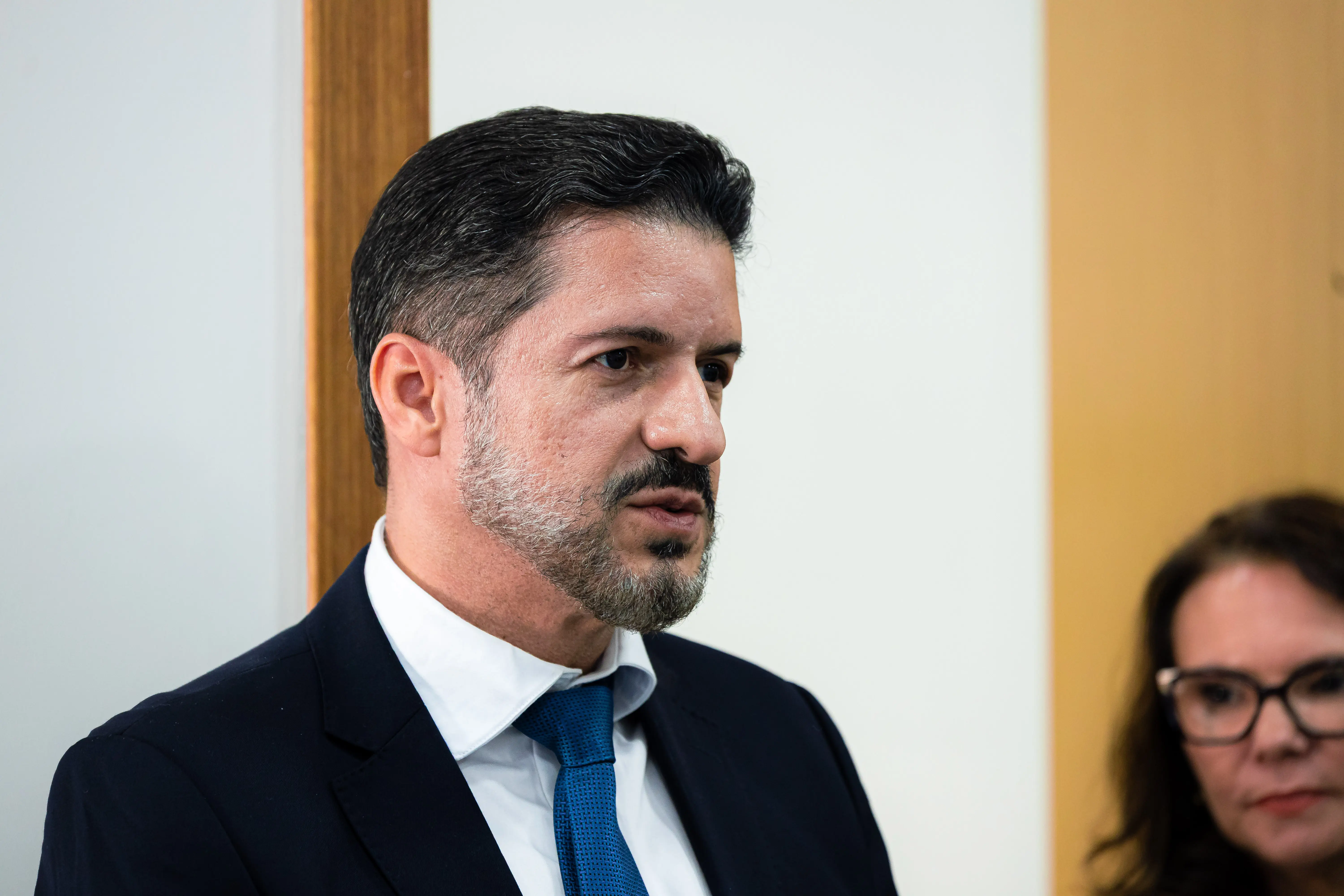 Homem de cabelos escuros, barba grisalha, terno azul-marinho, camisa branca e gravata azul, fala olhando para o lado. Ao fundo, parede branca e detalhe em madeira.