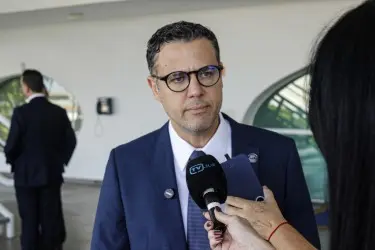 Homem de terno azul, camisa clara e óculos fala diante de microfone. . Ao fundo, espaço interno com vidro e profissionais desfocados.