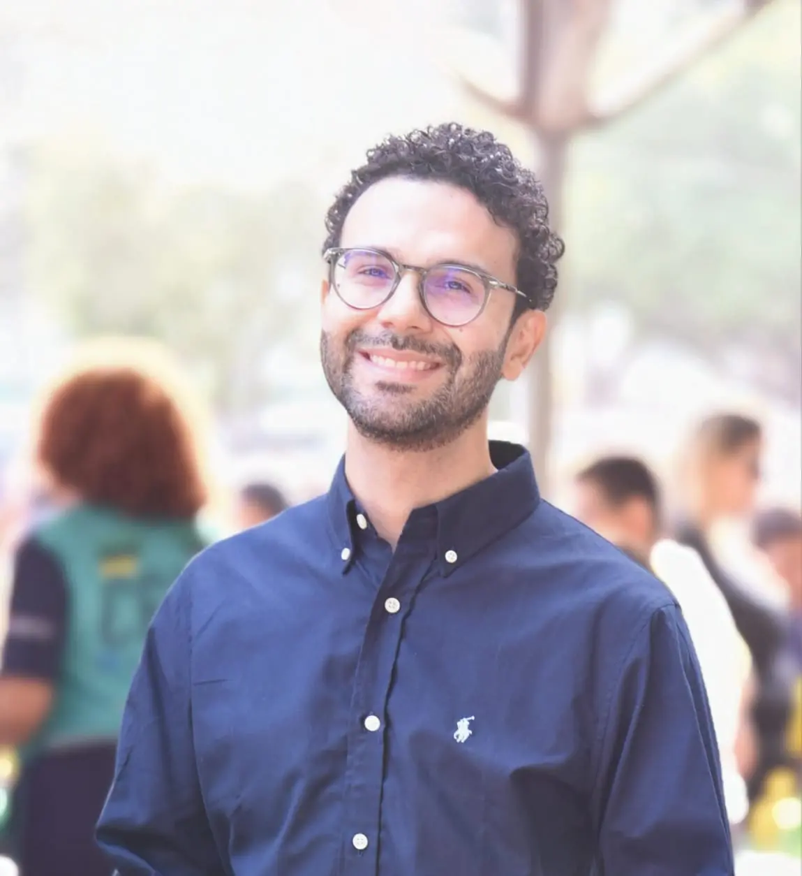 Homem sorrindo com barba curta, cabelos cacheados e óculos de grau. Veste camisa social azul-marinho. Está posicionado da cintura para cima, com fundo desfocado em ambiente externo.