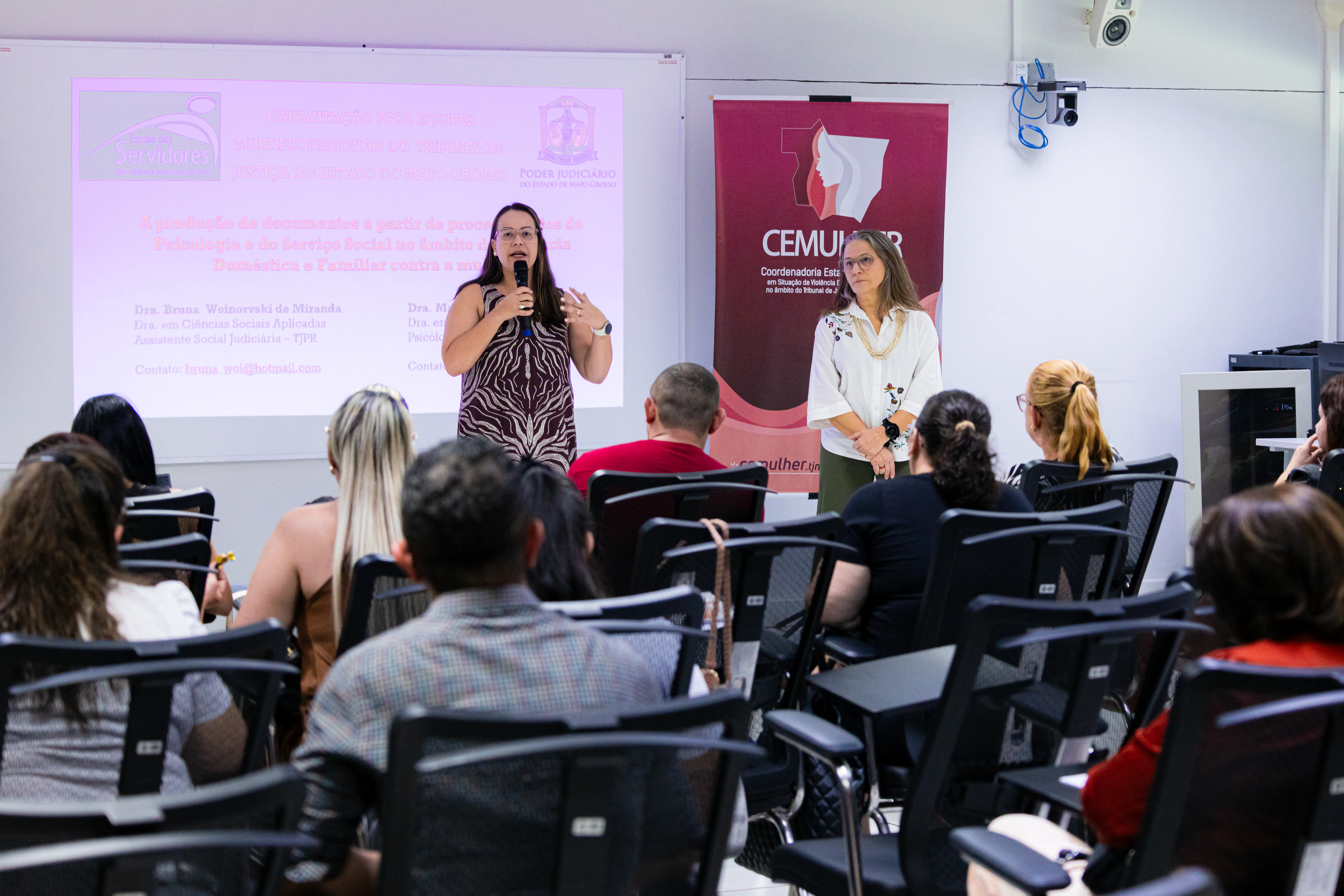 Duas mulheres palestram à frente de uma plateia sentada. À esquerda, uma delas fala ao microfone diante de uma tela de projeção; ao lado, outra observa em frente a um banner da CEMULHER.