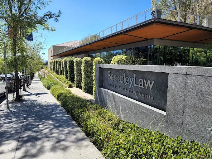 Foto horizontal que mostra a fachada da Berkeley Law da Universidade da Califórnia. É um muro de concreto com a placa da instituição, colunas de plantas e uma calçada arborizada sob um céu azul.