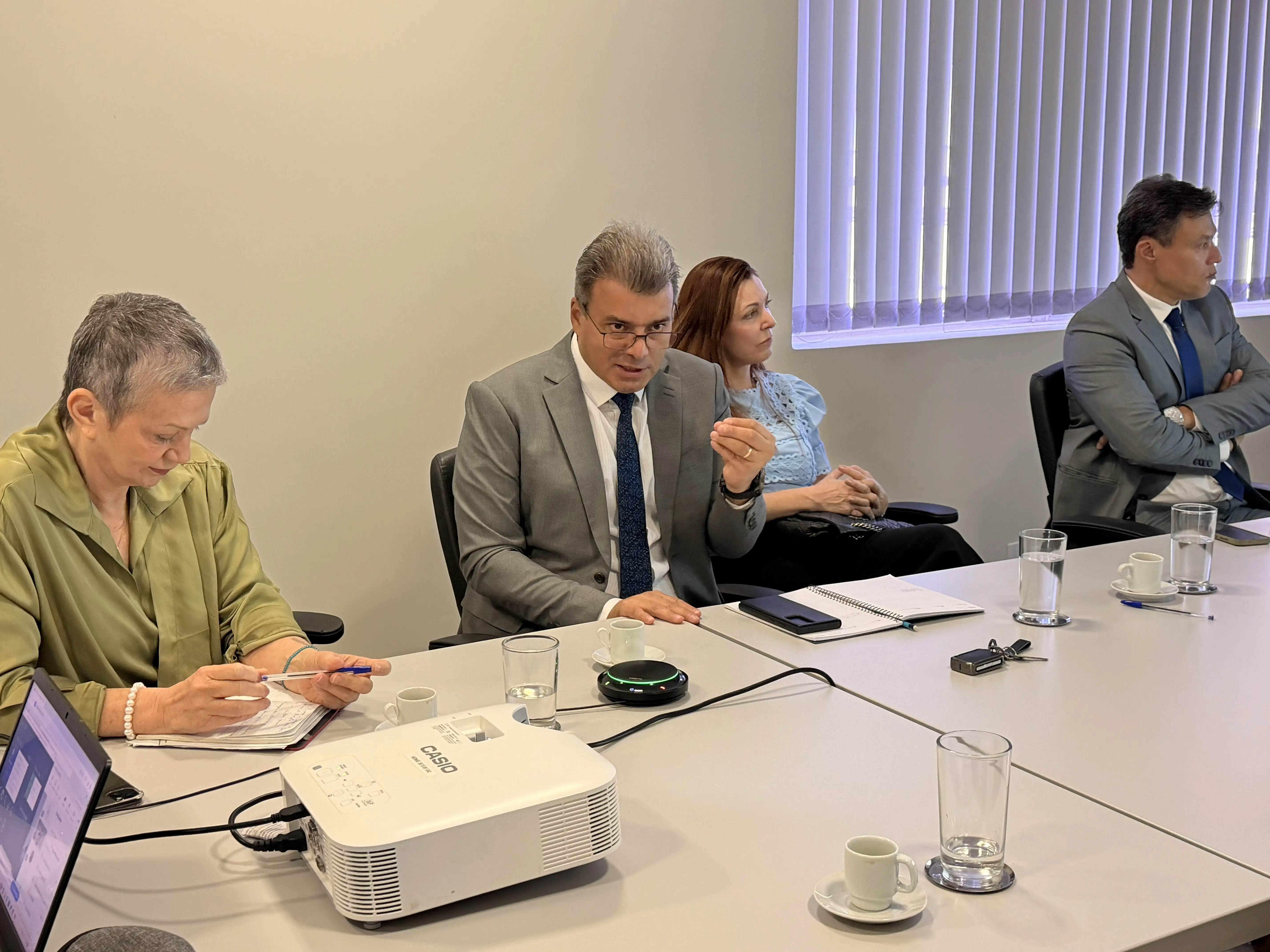Pessoas reunidas ao redor de uma mesa de reunião clara. Uma mulher de cabelo curto escreve em um bloco e um homem de terno cinza e óculos aponta o braço para a direita. Na mesa, há copos, xícaras de café, notebooks e um projetor branco. Ao fundo, uma janela com persianas.