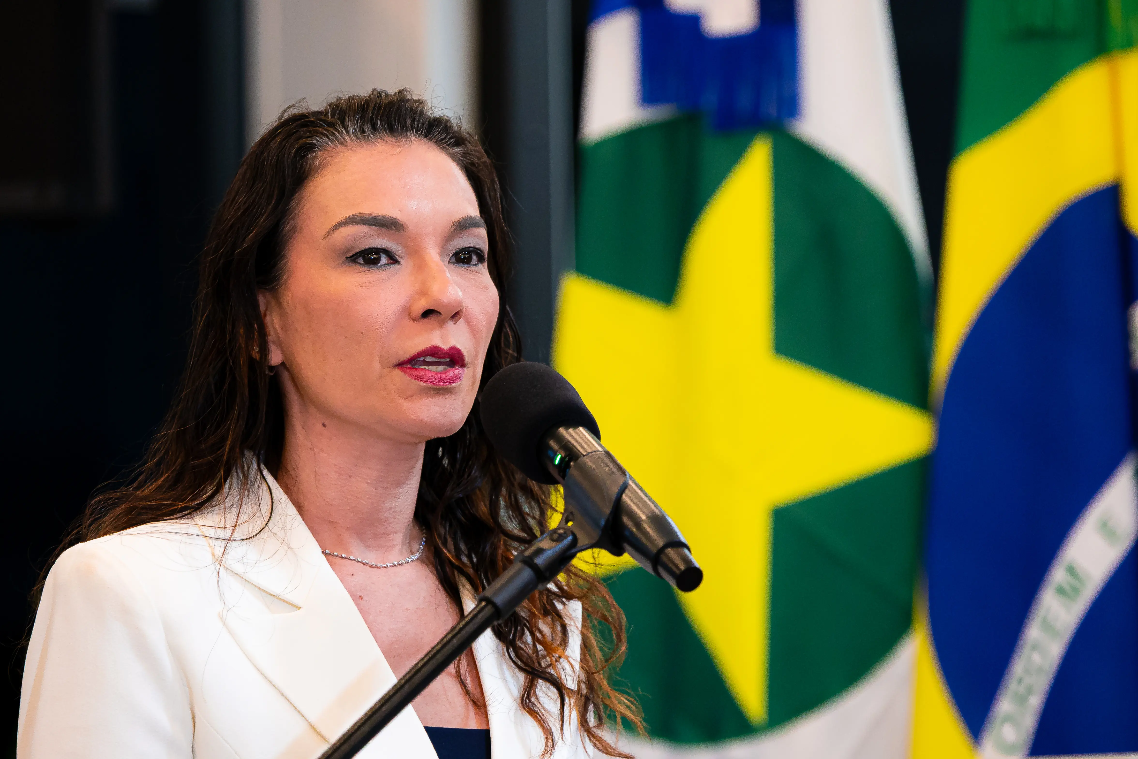Mulher de cabelos escuros ondulados veste blazer branco e colar prateado, fala ao microfone. Ao fundo, bandeira de Mato Grosso em destaque.