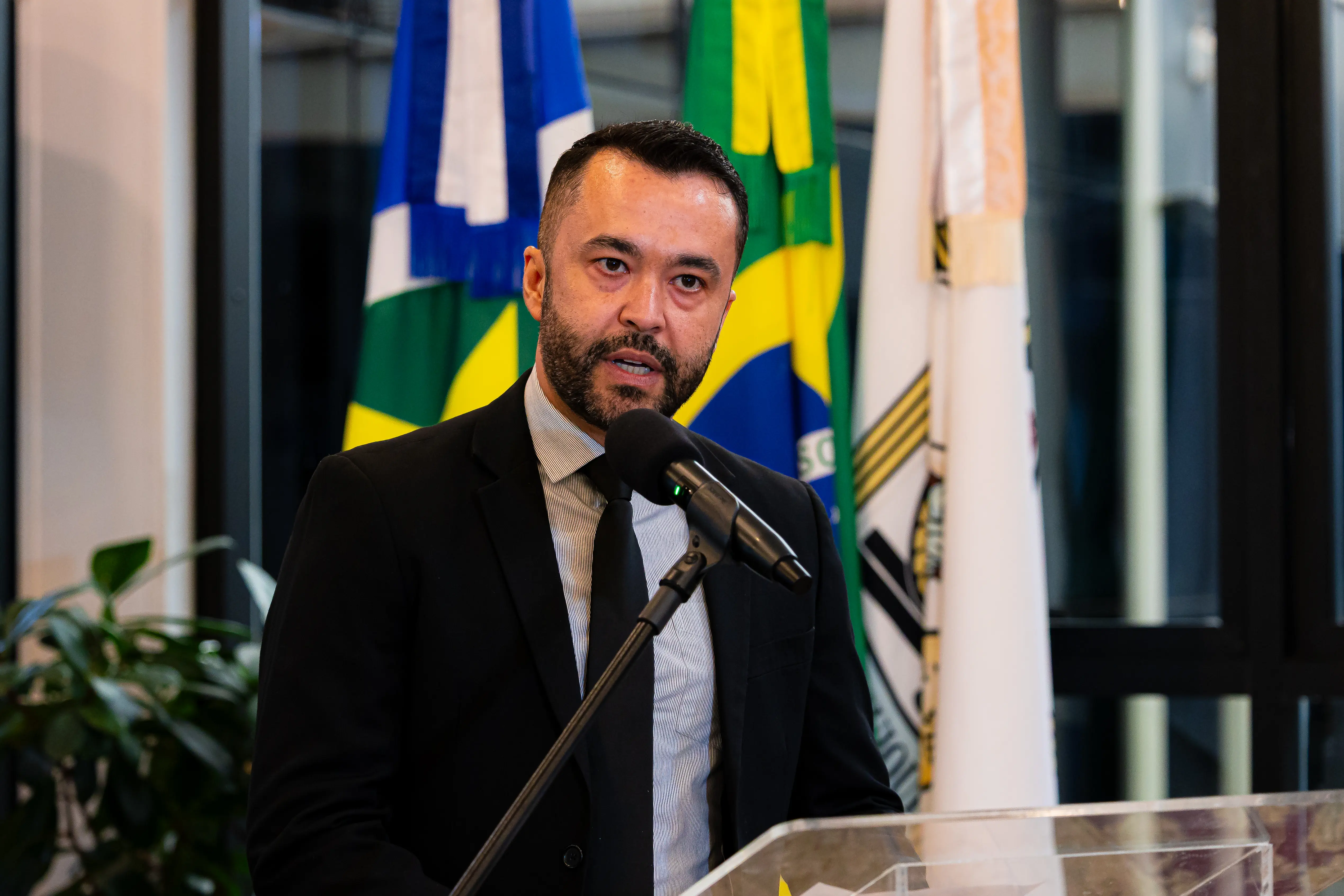Homem de cabelos escuros e barba, veste terno preto, camisa listrada e gravata preta, fala ao microfone. Ao fundo, bandeiras de Mato Grosso e do Brasil.