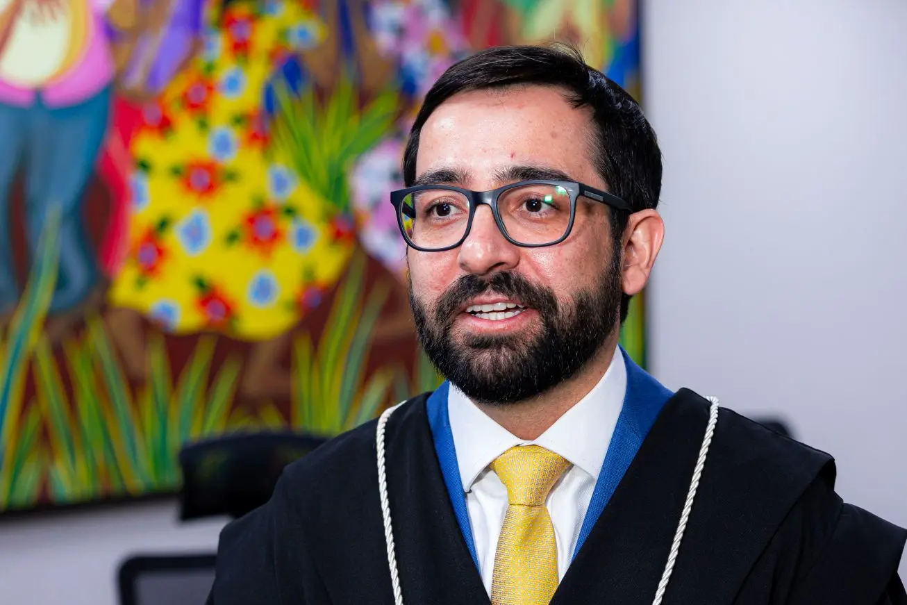Foto horizontal que mostra o juiz Eduardo Henrique Minosso do busto pra cima. Ele é um homem branco, de cabelo e barba castanhos escuro, usando óculos de grau e toga. Atrás dele há um quadro de pintura na parede.