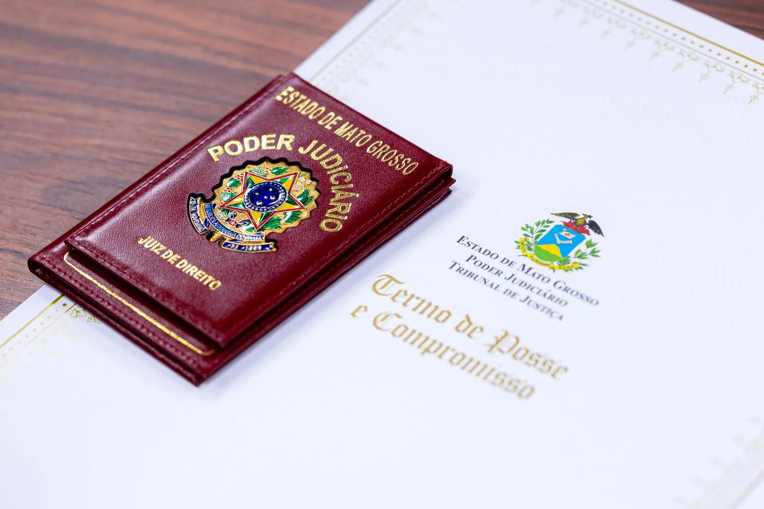 Foto horizontal que mostra a capa de um termo de posse e compromisso ao lado de uma carteira funcional de juiz de Direito dispostos sobre uma mesa. A carteira funcional é de couro vermelha, com o brasão do Poder Judiciário.
