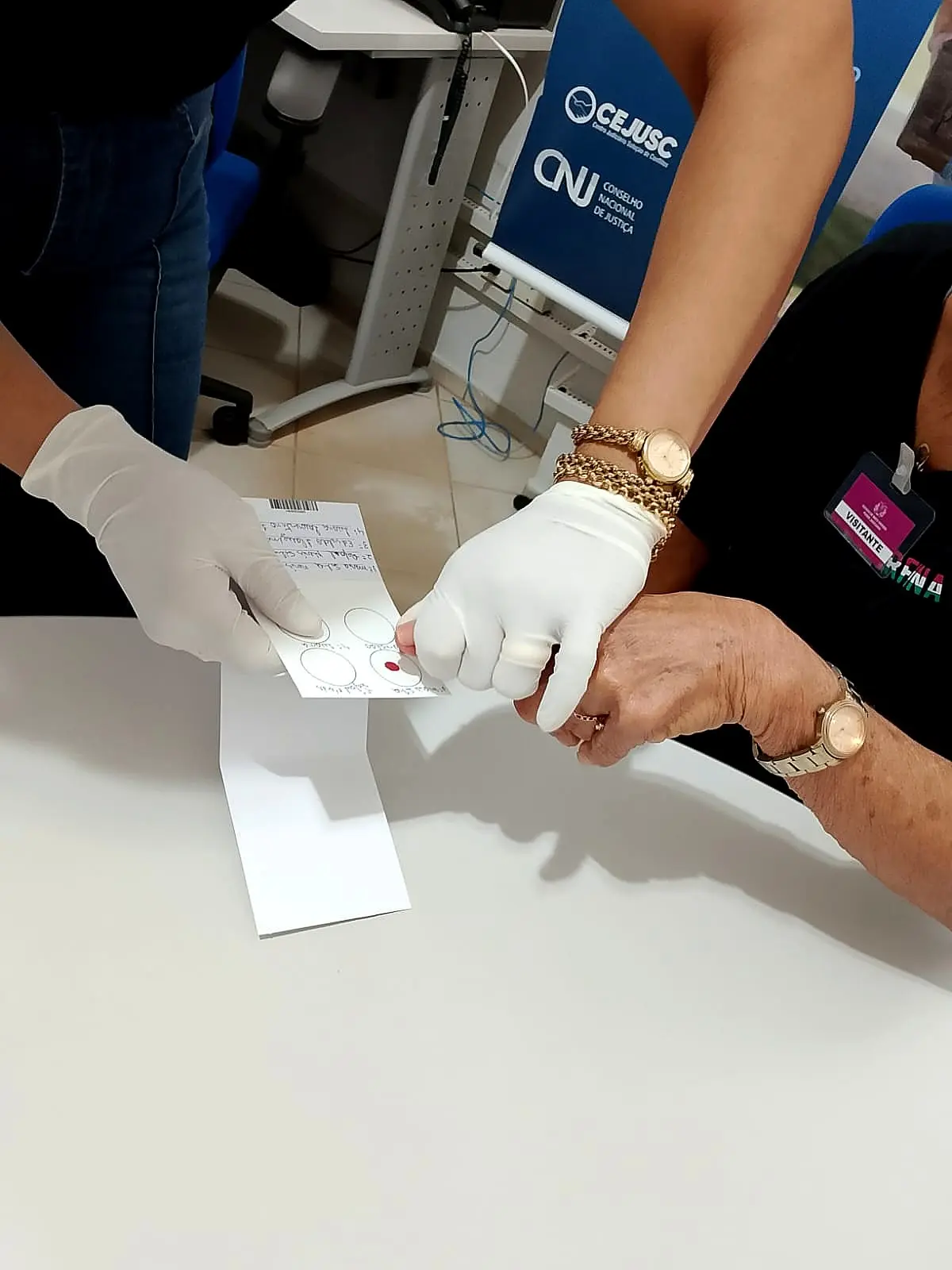 Mãos com luvas brancas coletam amostra de sangue em cartão de papel para exame de DNA, sobre mesa branca. Ao fundo, banners azuis do CEJUSC e do Conselho Nacional de Justiça (CNJ)