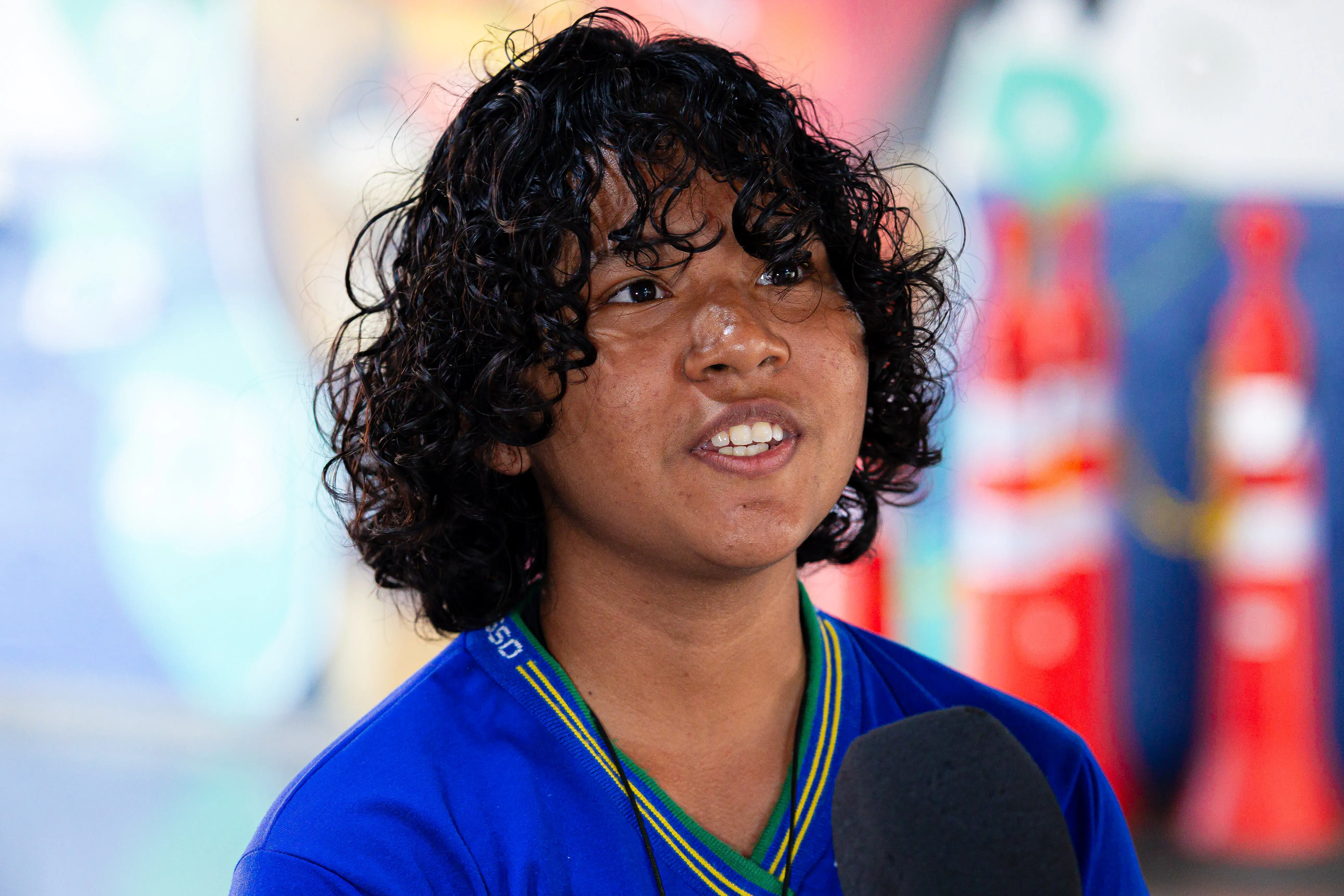 Estudante de cabelos cacheados escuros veste camiseta azul com gola listrada em verde, amarelo e azul, fala ao microfone. Ao fundo, mural colorido desfocado decora o ambiente escolar
