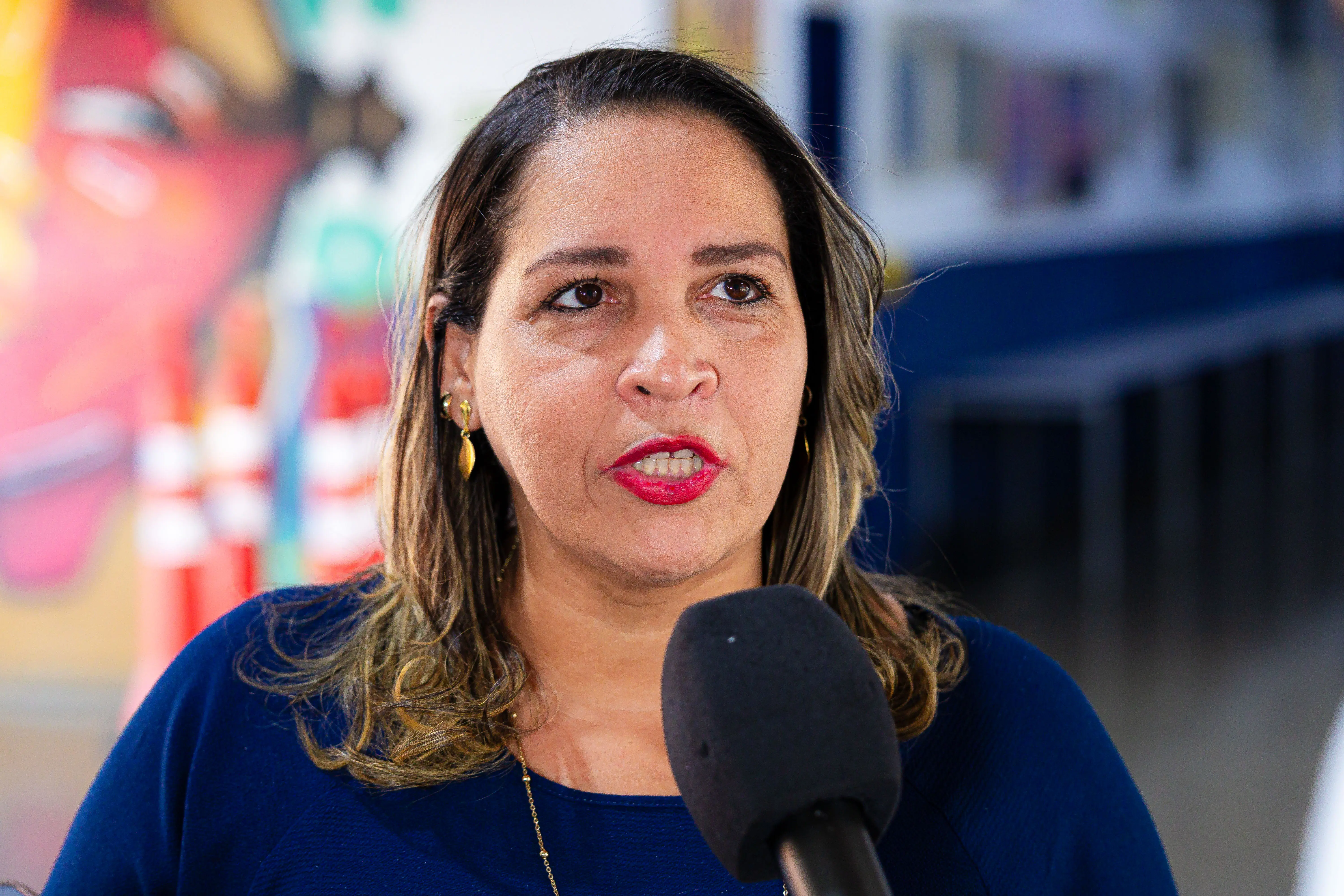 Mulher de cabelos escuros e batom vermelho, veste blusa azul, brincos e colar dourados, fala ao microfone. Ao fundo, mural colorido desfocado