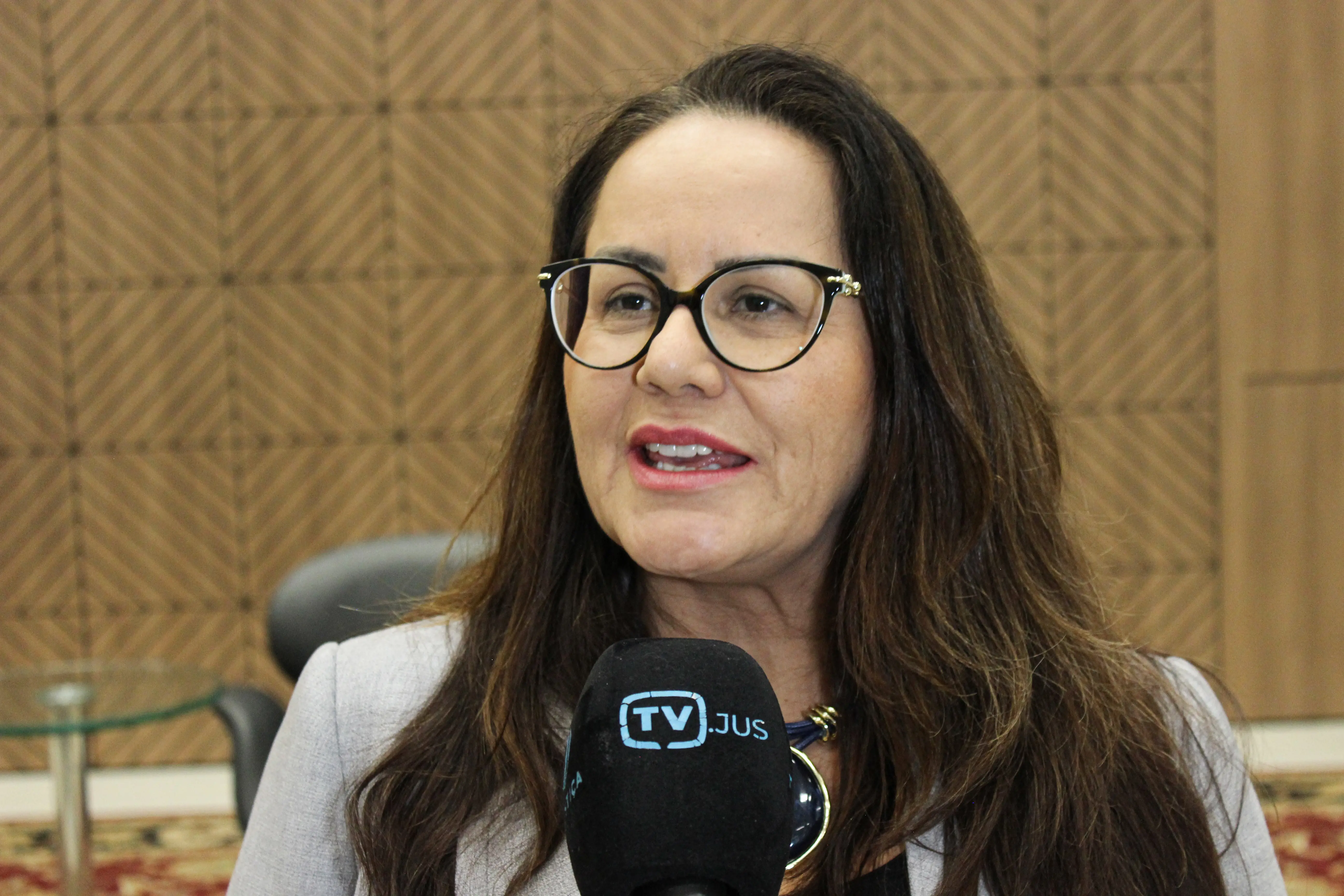 Mulher de cabelos escuros longos e óculos de armação preta falando próximo a um microfone com o logotipo 