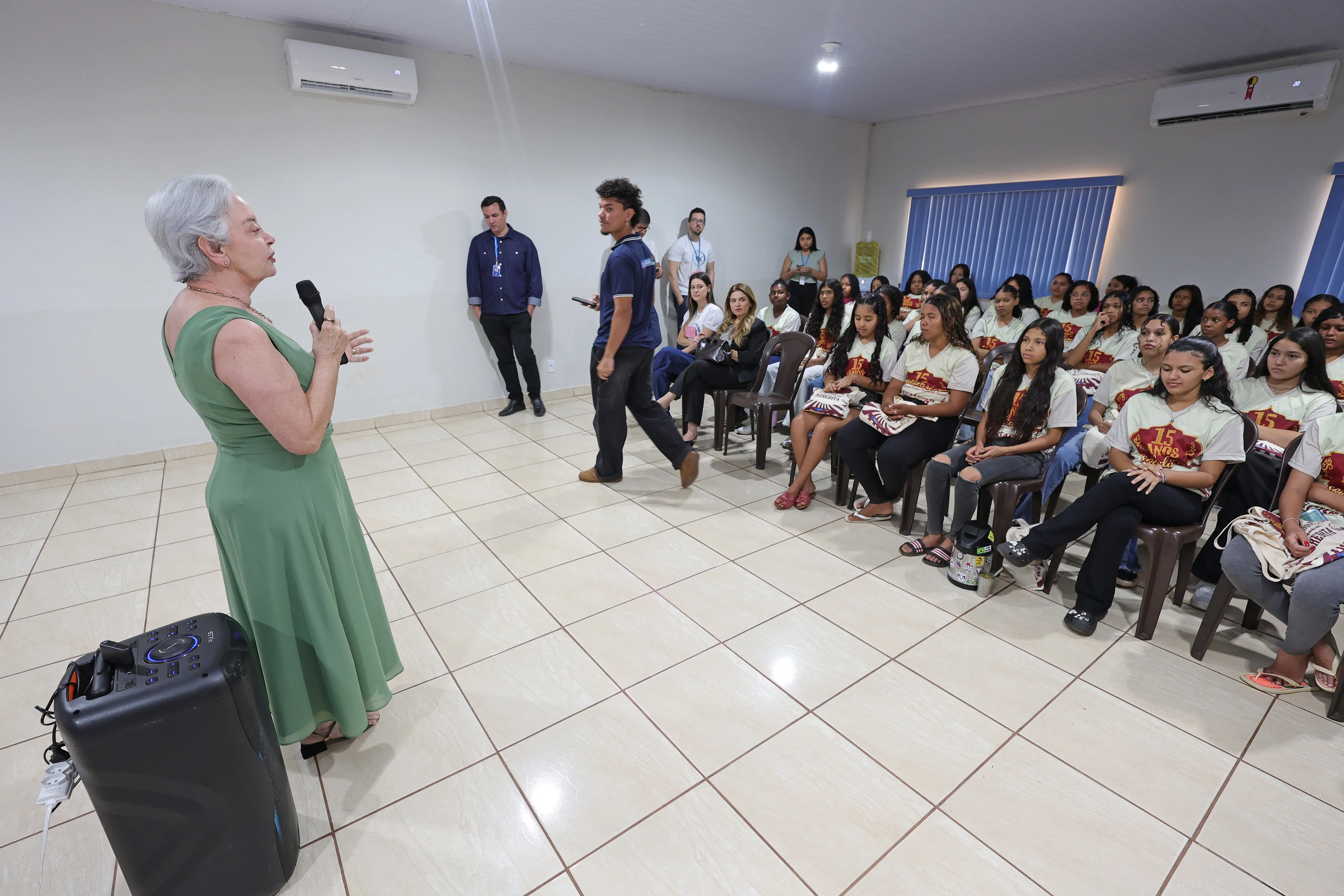 Mulher de vestido verde discursa ao microfone para uma plateia de jovens sentados em cadeiras. Uma caixa de som preta está no chão em primeiro plano.